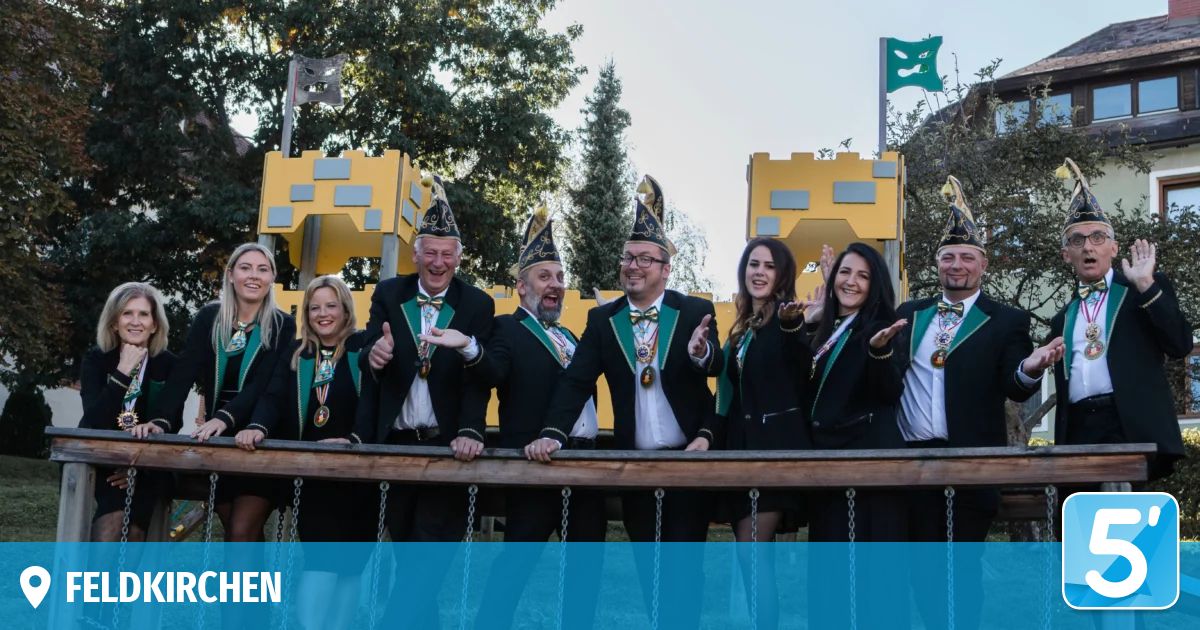 Eine Gruppe von Menschen in schwarzen Anzügen und grünen Hüten posiert für ein Foto vor einer burgartigen Struktur. Im Hintergrund befinden sich Bäume und Flaggen.