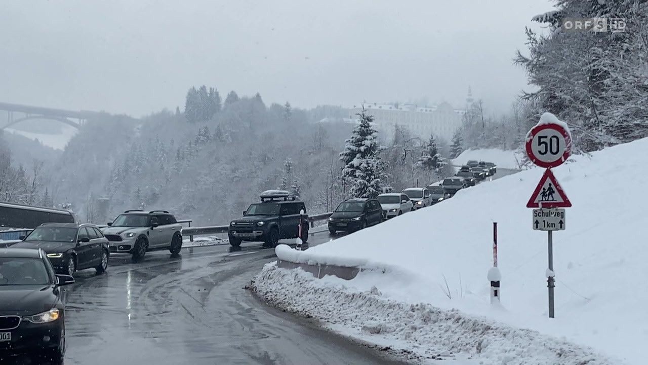 Eine Straße ist schneebedeckt und mehrere Autos stehen in einer Reihe, möglicherweise aufgrund eines Staus. Die Autos sind in einer Reihe am Straßenrand geparkt.