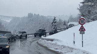 Eine Straße ist schneebedeckt und mehrere Autos stehen in einer Reihe, möglicherweise aufgrund eines Staus. Die Autos sind in einer Reihe am Straßenrand geparkt.