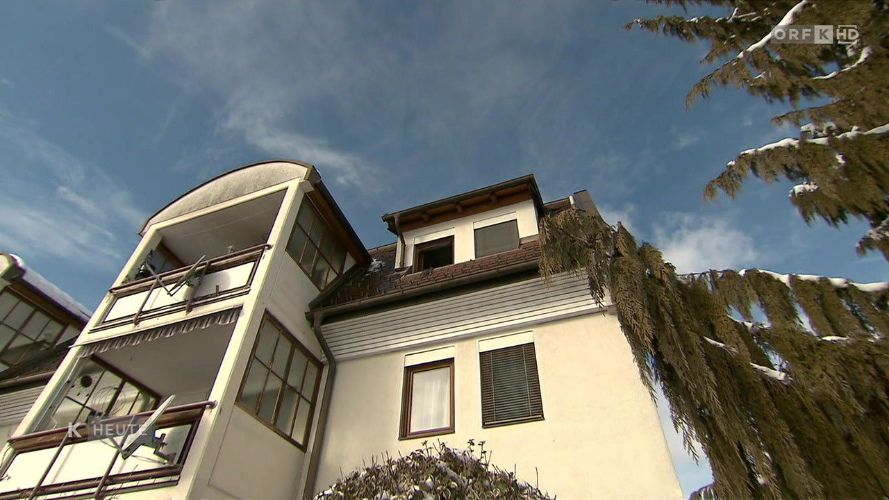 Ein zweistöckiges Gebäude mit einem braunen Dach und weißen Wänden ist abgebildet. Es hat mehrere Fenster, einige mit weißen Vorhängen. Das Gebäude verfügt über einen Balkon auf der linken Seite.