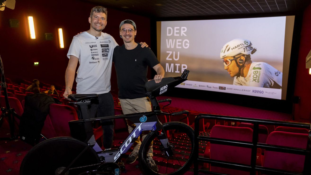 Zwei Männer stehen neben einem Scott-Fahrrad, einer in einem weißen T-Shirt, der andere in einem schwarzen T-Shirt. Sie sind in einem Kino mit einer Projektionsleinwand, die einen Radfahrer in einem weißen Helm zeigt.