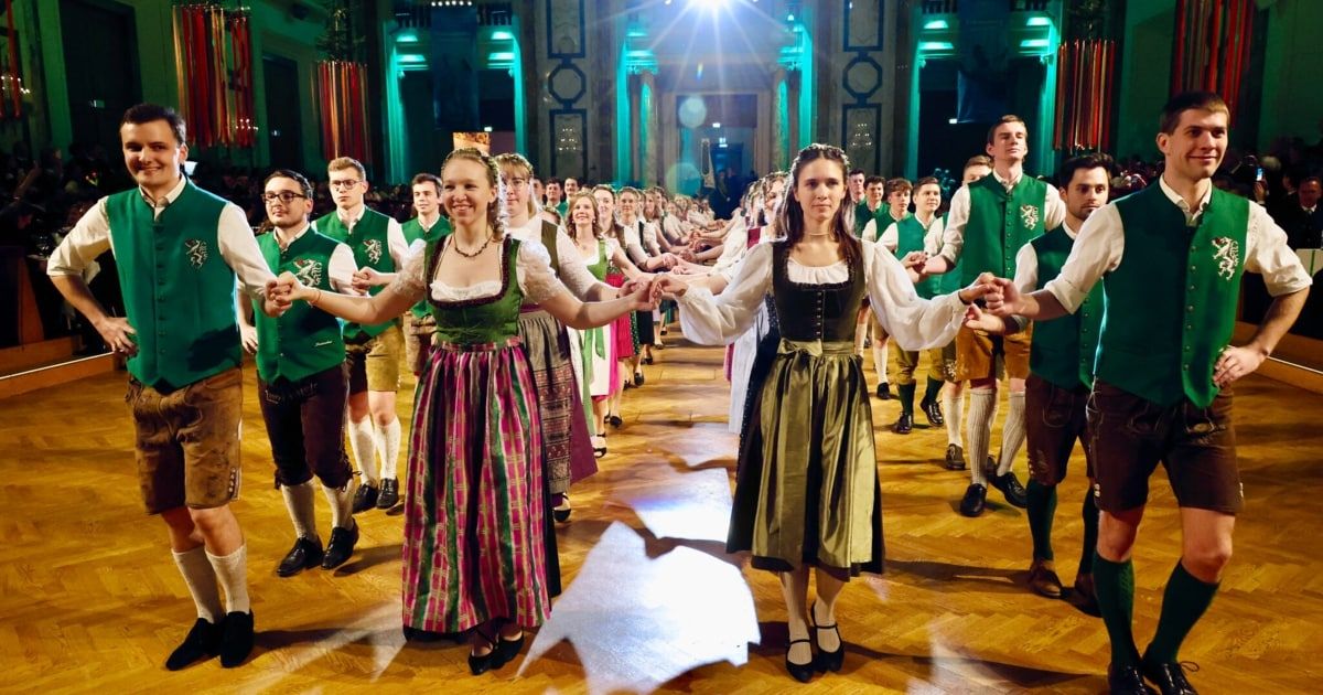 Eine Gruppe von Menschen in traditioneller deutscher Kleidung tanzt in einem großen Saal und hält dabei in einer Reihe die Hände.