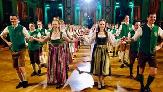 Eine Gruppe von Menschen in traditioneller deutscher Kleidung tanzt in einem großen Saal und hält dabei in einer Reihe die Hände.