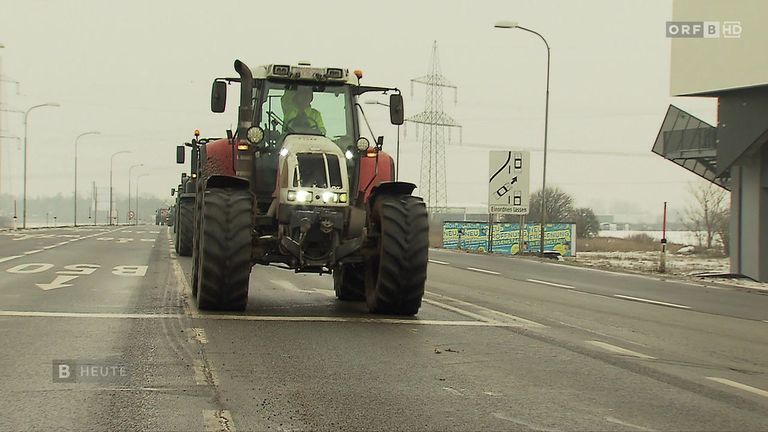 Ein Traktor mit eingeschalteten Scheinwerfern fährt auf einer Straße mit weißen Markierungen und einem Geschwindigkeitsschild, begleitet von anderen Fahrzeugen.