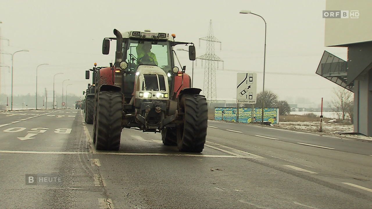 Ein Traktor mit eingeschalteten Scheinwerfern fährt auf einer Straße mit weißen Markierungen und einem Geschwindigkeitsschild, begleitet von anderen Fahrzeugen.