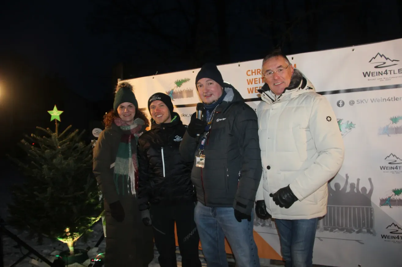 Vier Personen stehen bei Nacht vor einem Weihnachtsbaum und tragen Winterkleidung. Ein Mann rechts hält ein Mikrofon, und ein Schild hinter ihnen zeigt 'Weihnachten' auf Deutsch.
