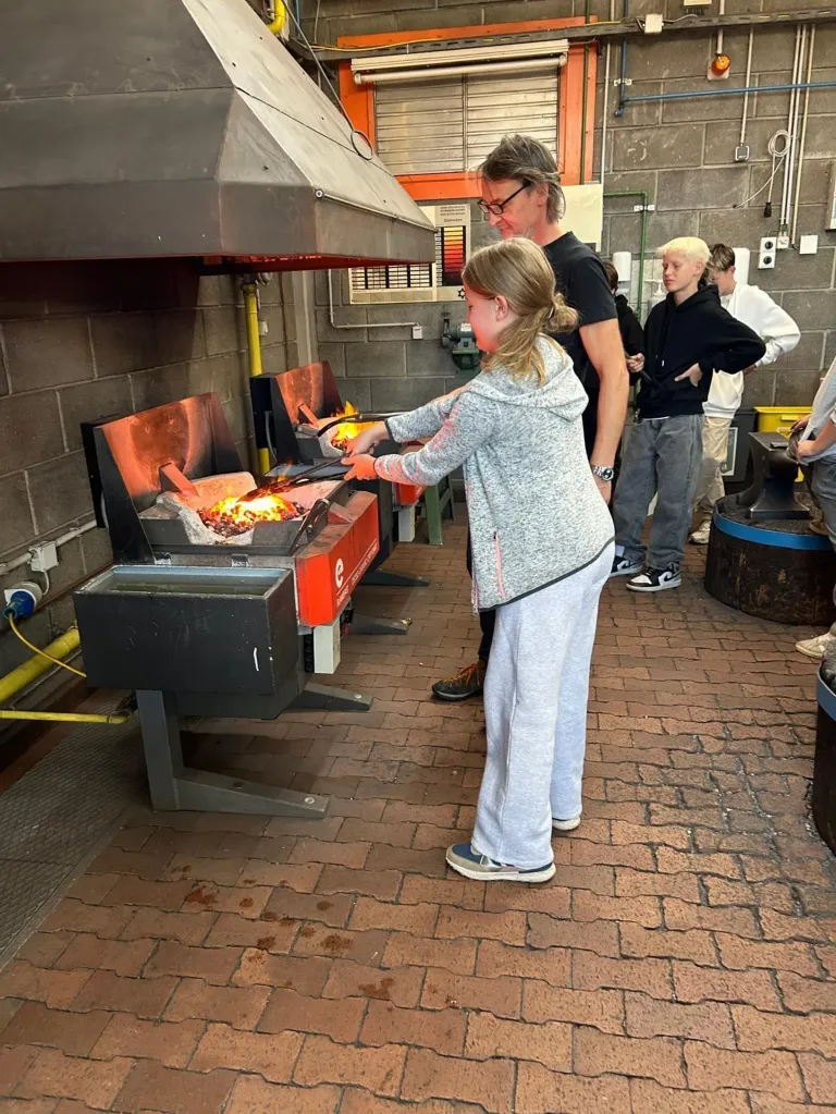 Ein Mädchen mit einer grauen Kapuzenjacke arbeitet in einer Werkstatt an einem Metallobjekt. Sie benutzt ein Paar Zangen. Hinter ihr lächelt ein Mann und beobachtet sie. Vor ihr befinden sich zwei Metallobjekte. Es stehen noch andere Leute herum.