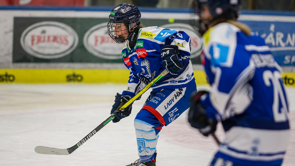 Zwei weibliche Hockeyspielerinnen in blauen Trikots skaten auf einer Eisbahn. Eine Spielerin schaut die andere an. Hinter ihnen ist ein unscharfer Hintergrund mit Sponsorenlogos.