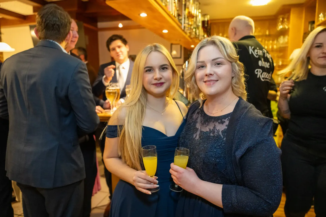 Zwei Frauen in Abendkleidung lächeln für die Kamera auf einer Party. Sie halten Gläser mit Orangensaft. Im Hintergrund sind ein Mann im Anzug und ein Barkeeper zu sehen.