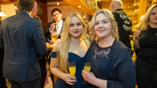 Zwei Frauen in Abendkleidung lächeln für die Kamera auf einer Party. Sie halten Gläser mit Orangensaft. Im Hintergrund sind ein Mann im Anzug und ein Barkeeper zu sehen.