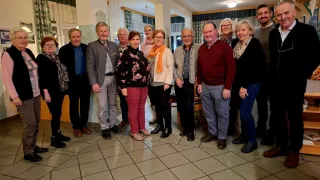 Eine Gruppe von Menschen in formeller Kleidung posiert für ein Foto in einem Raum mit gefliestem Boden. Einige lächeln, während andere direkt in die Kamera blicken. Der Raum hat weiße Wände und Decke, und es hängen Vorhänge an den Fenstern.