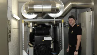 Ein Mann in einem schwarzen Hemd steht neben einer großen schwarzen und silbernen Industriemaschine mit silbernen Rohren. Er lächelt und schaut nach vorne.