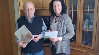 Ein älterer Mann und eine Frau stehen vor einem Holzschrank und halten Bücher und Umschläge in den Händen.