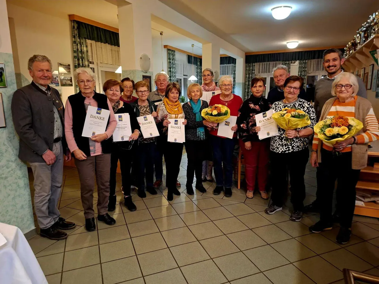 Eine Gruppe älterer Menschen, überwiegend Frauen, steht in einem Raum und hält Urkunden und Blumen. Sie sind in Winterkleidung gekleidet und stehen auf einem gefliesten Boden.