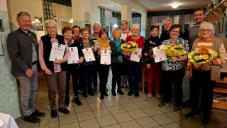 Eine Gruppe älterer Menschen, überwiegend Frauen, steht in einem Raum und hält Urkunden und Blumen. Sie sind in Winterkleidung gekleidet und stehen auf einem gefliesten Boden.