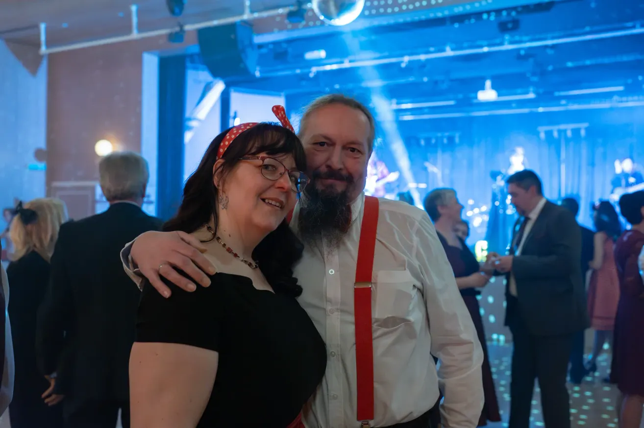 Ein Mann und eine Frau stehen bei einer Veranstaltung eng beieinander. Die Frau trägt eine Brille und eine rote Kopfschleife. Der Mann hat einen Bart und rote Träger. Andere Leute sind im Hintergrund.