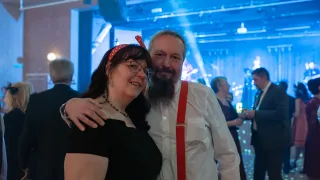 Ein Mann und eine Frau stehen bei einer Veranstaltung eng beieinander. Die Frau trägt eine Brille und eine rote Kopfschleife. Der Mann hat einen Bart und rote Träger. Andere Leute sind im Hintergrund.