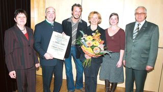 Eine Gruppe von Menschen posiert für ein Foto. Ein Mann hält ein Zertifikat und ein anderer eine Blumenstrauß. Sie stehen vor einer Wand.