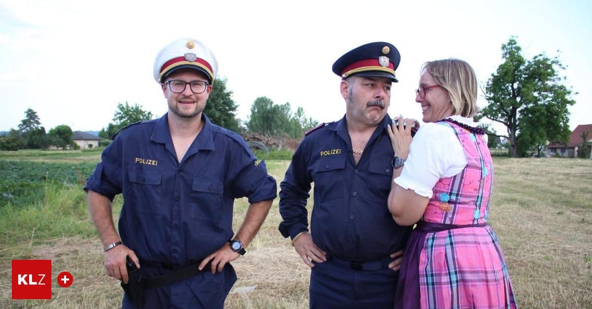 Zwei Polizisten und eine Frau stehen auf einem Feld, lächeln und posieren für ein Foto. Die Polizisten tragen marineblaue Uniformen und Mützen. Die Frau trägt ein farbenfrohes Kleid.