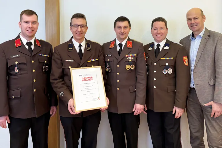 Vier Männer in Militäruniformen, einschließlich Medaillen, stehen zusammen und halten ein Zertifikat. Sie lächeln und stehen vor einer weißen Wand.