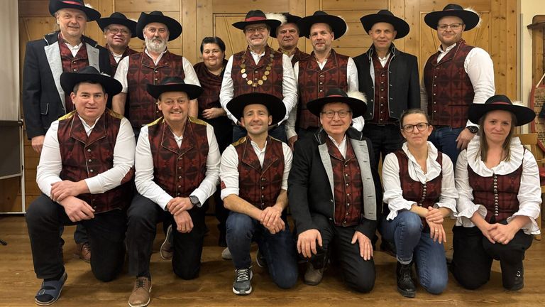 Eine Gruppe von Menschen in traditionellen Kostümen, darunter Männer in schwarzen Hüten und Westen und Frauen in langen Kleidern, posieren für ein Foto. Sie stehen vor einer Holzwand.