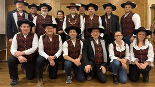 Eine Gruppe von Menschen in traditionellen Kostümen, darunter Männer in schwarzen Hüten und Westen und Frauen in langen Kleidern, posieren für ein Foto. Sie stehen vor einer Holzwand.