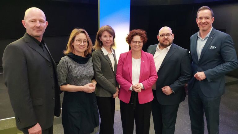 Fünf Personen, vier Frauen und ein Mann, stehen zusammen und lächeln für ein Foto. Sie sind feierlich gekleidet, wobei die Frau in der Mitte eine rosafarbene Jacke trägt.