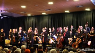 Ein Symphonieorchester spielt auf der Bühne mit einem Dirigenten vor einem Publikum, einige spielen Violinen, Celli und Trompeten.