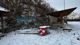 Eine verschneite Szene mit einem roten Fass mit Puzzle-Design, zwei grauen Fässern, einem hölzernen Pavillon und einem verschneiten Hügel mit Bäumen.