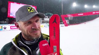 Ein Skifahrer mit Atomic-Skiern posiert im Schnee und hält ein Mikrofon. Er trägt eine graue Mütze mit einem gelben X und eine grüne Jacke. Hinter ihm ist eine rote Barriere mit dem Wort Audi zu sehen.