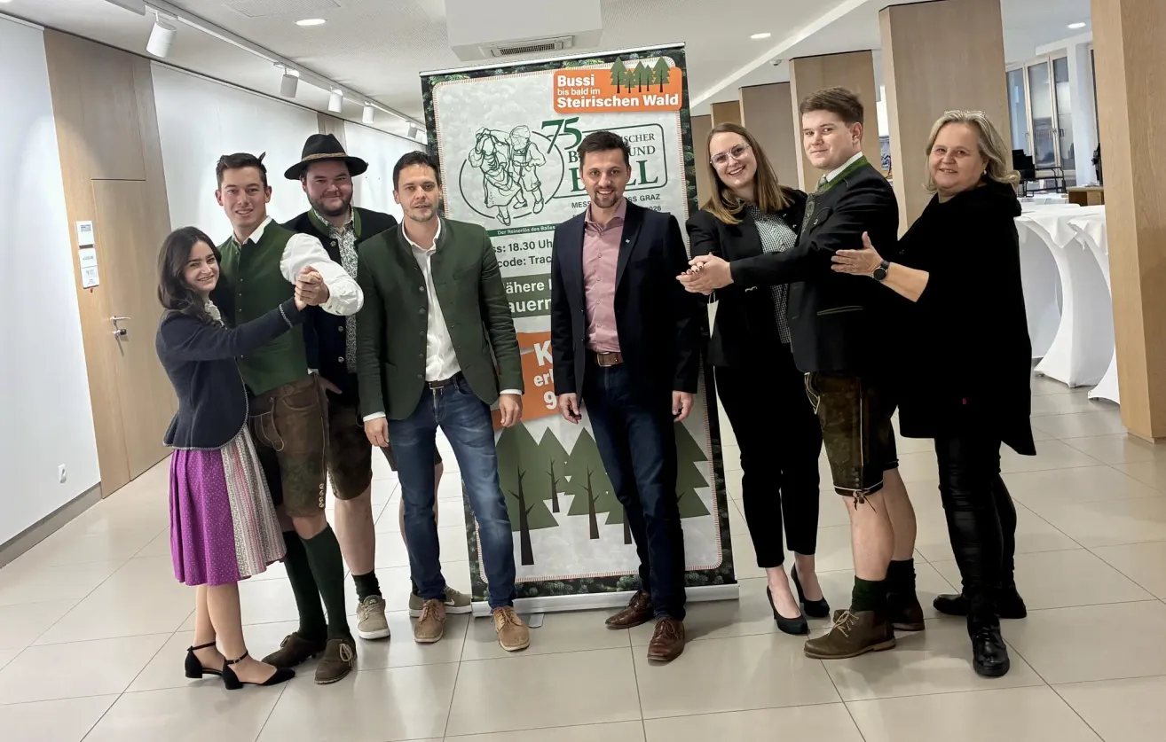 Eine Gruppe von Menschen steht vor einem Plakat zum 75-jährigen Jubiläum von Bussi im Steirischen Wald und trägt traditionelle Kleidung.