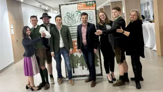 Eine Gruppe von Menschen steht vor einem Plakat zum 75-jährigen Jubiläum von Bussi im Steirischen Wald und trägt traditionelle Kleidung.