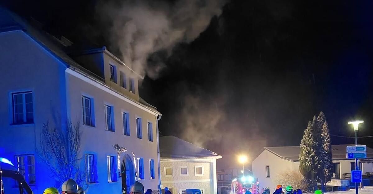 In der Nacht steigt Rauch vom Dach eines Gebäudes mit mehreren Fenstern auf. Ein Feuerwehrauto ist in der Nähe geparkt und seine Lichter sind an.