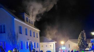 In der Nacht steigt Rauch vom Dach eines Gebäudes mit mehreren Fenstern auf. Ein Feuerwehrauto ist in der Nähe geparkt und seine Lichter sind an.