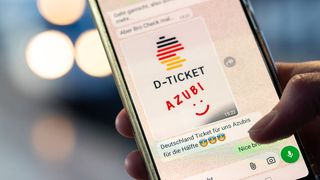 Ein Smartphone zeigt eine Nachricht mit dem Text D-TICKET 24, einem Emoji mit einem lächelnden Gesicht und einer Zeitangabe von 15:22. Die Nachricht ist für ein Ticket für Deutschland. Der Hintergrund ist unscharf mit Bokeh-Lichtern.