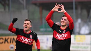 Zwei Männer in Sporttrikots feiern auf einem Feld. Einer bildet mit den Händen eine Herzform, der andere schreit mit offenem Mund. Der Hintergrund ist unscharf.