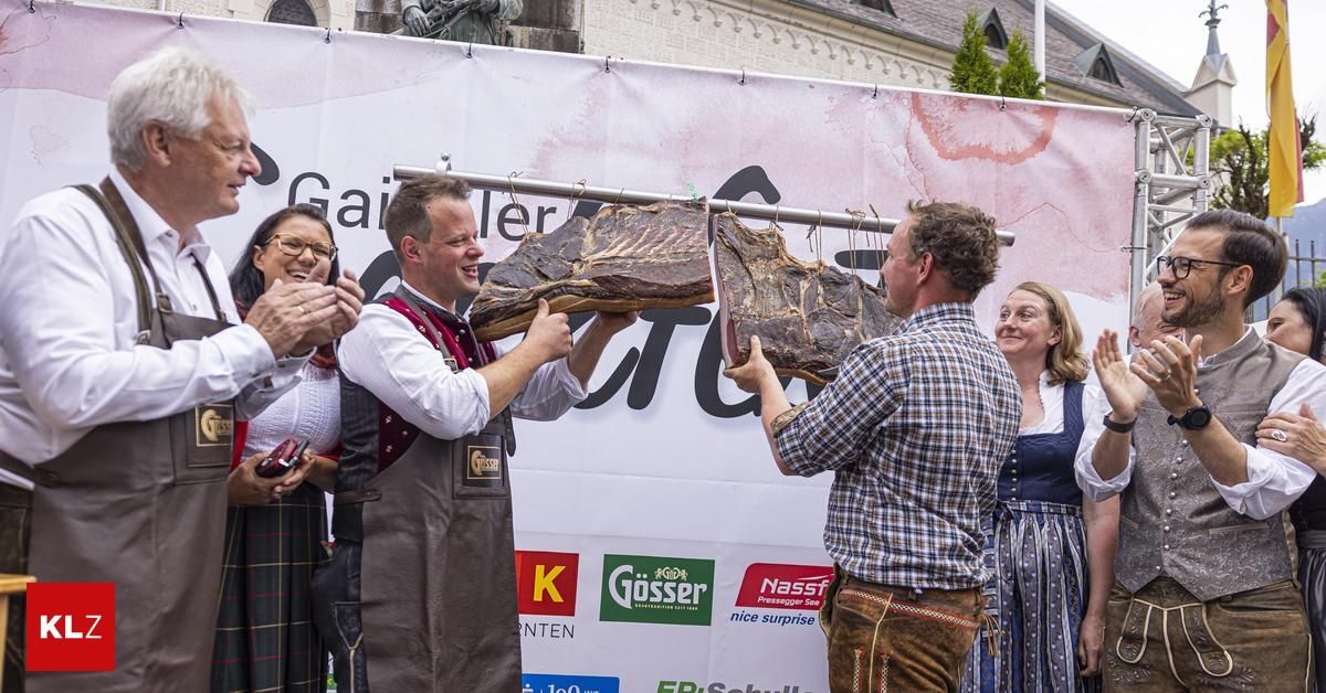 Zwei Männer präsentieren ein großes Stück Fleisch bei einem Festival. Ein Mann hält das Fleisch hoch, während der andere darauf zeigt. Hinter ihnen klatschen und lächeln die Leute. Ein Banner mit Text im Hintergrund.