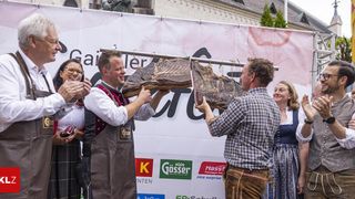 Zwei Männer präsentieren ein großes Stück Fleisch bei einem Festival. Ein Mann hält das Fleisch hoch, während der andere darauf zeigt. Hinter ihnen klatschen und lächeln die Leute. Ein Banner mit Text im Hintergrund.