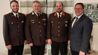 Vier Männer in formellen Uniformen stehen zusammen. Die beiden in der Mitte tragen militärische Kleidung mit Medaillen, während der Mann auf der rechten Seite eine Brille und einen Anzug trägt.