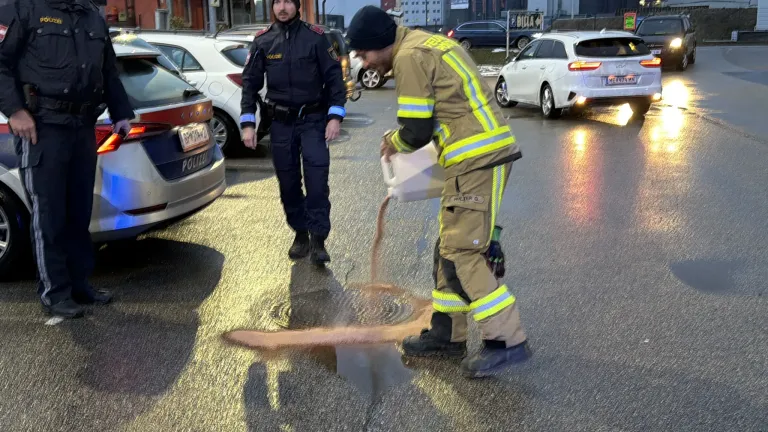 Ein Feuerwehrmann gießt Wasser in einen Schachtdeckel, während ein Polizeibeamter in der Nähe steht. Dahinter sind mehrere Autos geparkt, und Gebäude sind in der Ferne zu sehen.