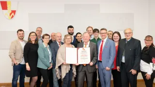 Eine Gruppe von Menschen, darunter Männer und Frauen, posiert für ein Foto. Eine Frau hält eine Urkunde, während ein Mann einen Ordner in der Hand hat. Dahinter ist eine weiße Wand mit geometrischen Mustern zu sehen.