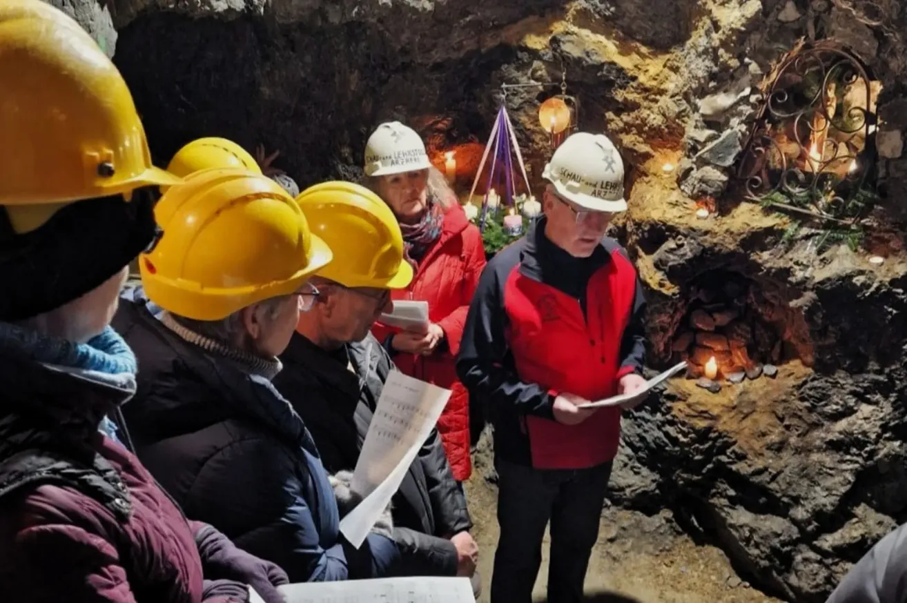 Eine Gruppe von Menschen mit gelben Helmen versammelt sich in einer Höhle. Sie scheinen einem Mann in einer roten Jacke zuzuhören, der von einem Stück Papier liest.