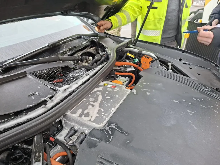 Eine Person in einer Sicherheitsweste inspiziert einen Automotor mit offenem Motorhaube, der freiliegende Drähte und einen Batteriefach zeigt.