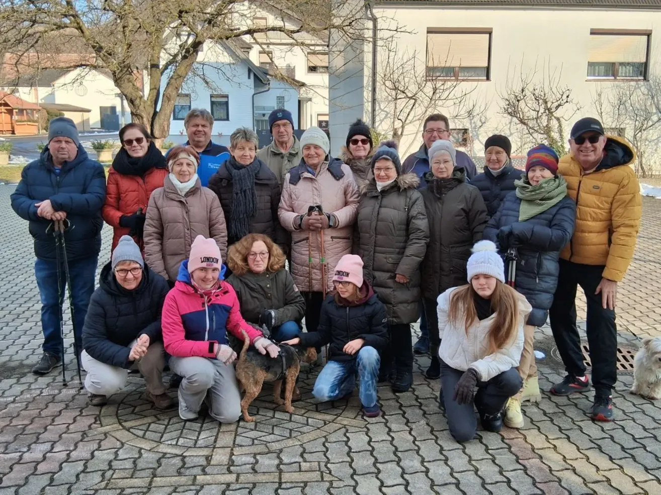 Eine Gruppe von Menschen in Winterkleidung posiert mit einem Hund vor einem Gebäude mit Bäumen.