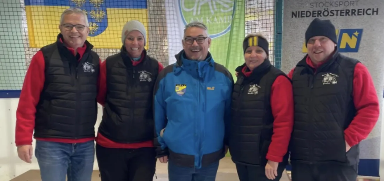 Drei Personen stehen eng beieinander für ein Foto. Ein Mann in der Mitte trägt eine Brille und eine blaue Jacke. Die beiden Frauen auf beiden Seiten tragen schwarze Westen. Hinter ihnen ist ein grüner Banner.