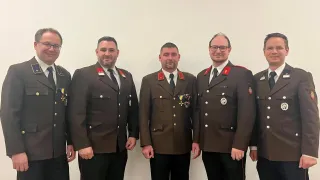 Drei Männer in Uniform stehen nebeneinander und lächeln für ein Foto. Sie tragen Medaillen und haben einen roten Rand an ihren Uniformen.