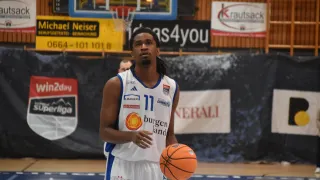 Ein Basketballspieler steht auf dem Spielfeld, hält einen orangefarbenen Ball in der Hand, trägt ein weißes Trikot mit der Nummer 17 und dem Wort Burgenland. Dahinter befindet sich ein Basketballkorb.