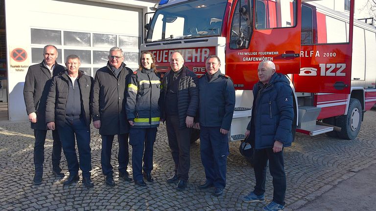 Sechs Personen stehen vor einem Feuerwehrauto. Einer trägt eine Feuerwehruniform mit reflektierenden Streifen. Sie stehen vor einer Garage.