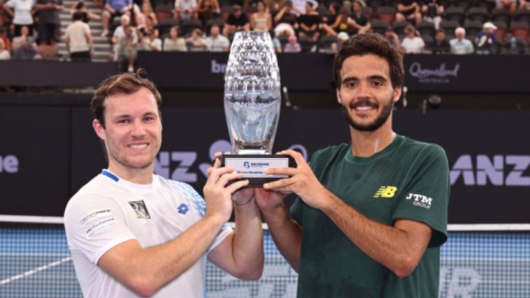 Zwei Männer halten eine Trophäe mit einem Brisbane-Logo, lächeln und posieren für ein Foto vor einer Menge.