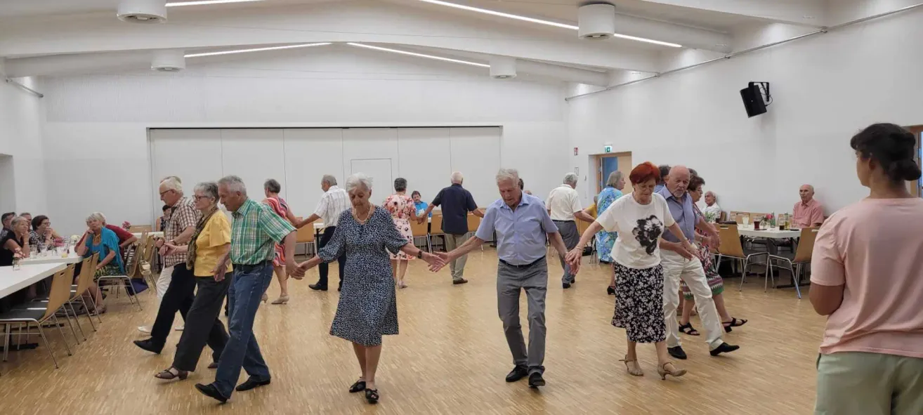 Mehrere ältere Paare tanzen gemeinsam in einem großen Raum. Sie alle lächeln und scheinen sich zu amüsieren. Der Raum hat weiße Wände und einen Holzboden.